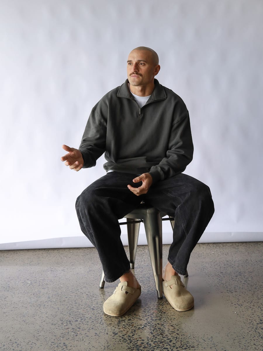 Sam Powell-Pepper seated on a stool in front of a white backdrop, mid-sentence.